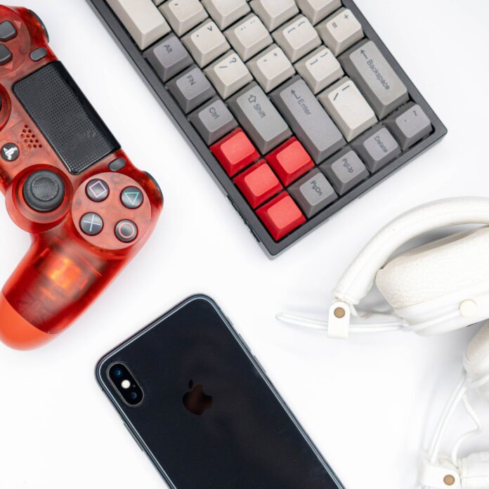 Roter Playstation-Controller neben Tastatur, weißen Kopfhörern und einem schwarzen Smartphone.